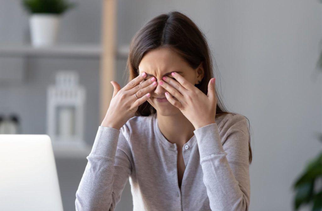 A woman inside the office sitting at a desk rubbing her eyes with her fingers due to eye problems.