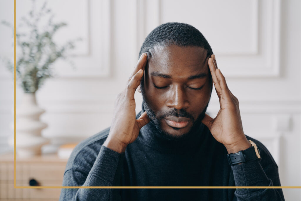 A man sitting indoors with his eyes closed, pressing his fingers to his temples, experiencing a migraine.